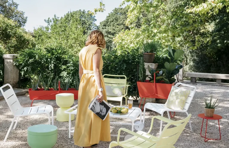 Frau in gelbem Kleid steht in einem Garten, umgeben von bunten Möbeln von Fermob und Pflanzen.